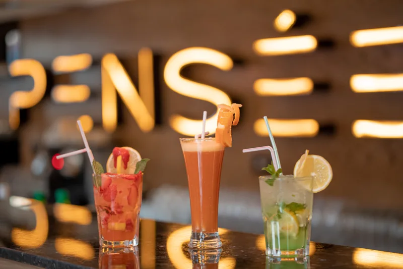 Trois boissons garnies de fruits devant l’enseigne lumineuse du restaurant Pensée à Marrakech